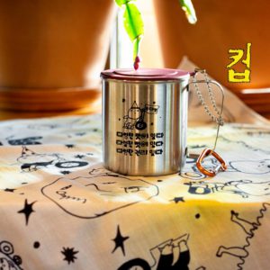 따뜻한 햇살을 받는 스테인리스 컵 클로즈업. 컵에는 그림과 글이 다음과 같이 프린트되어 있다. 들다방이라 쓰인 깃발을 휠체어에 달고 파티 모자를 쓴 채 나아가는 사람 그림. 하단에는 동글동글 가리런한 손글씨체로 ‘다양한 맛이 있다, 다양한 멋이 있다, 다양한 권리 있다 들다방’. 컵의 바닥에는 들다방 노랑 손수건이 깔려 있다.